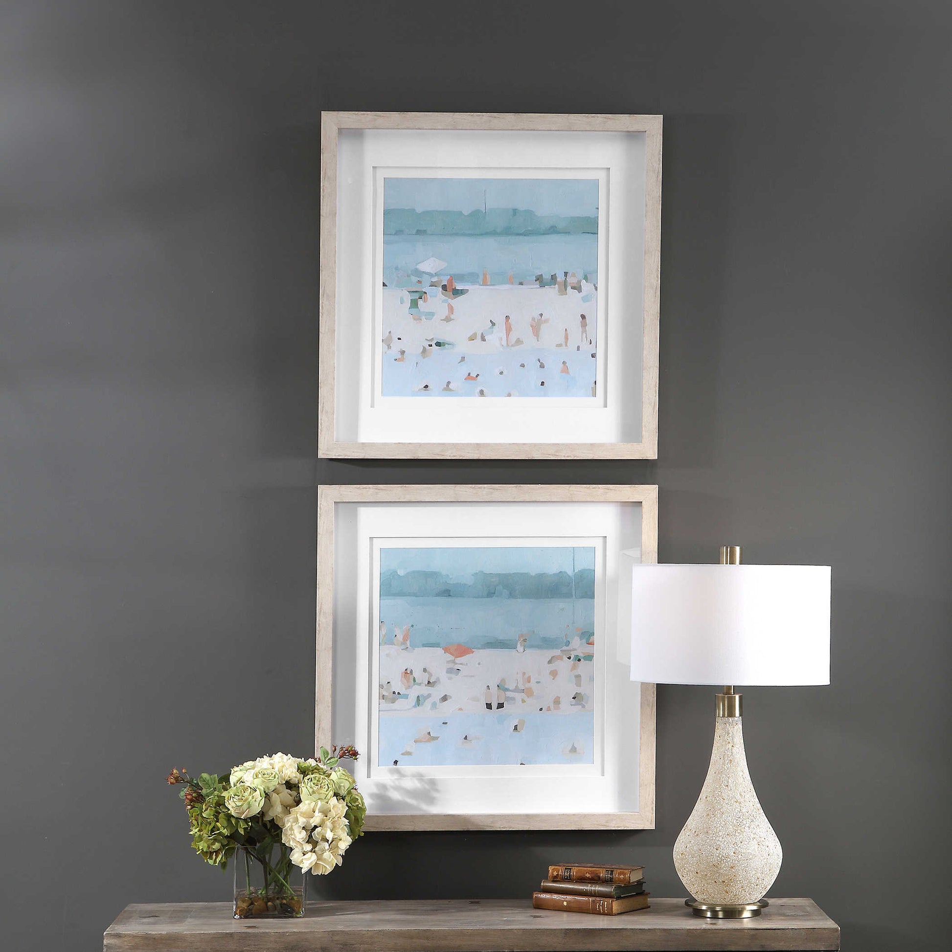 Stacked small art depicting a beach coastal scene on top of a console with a lamp, books, and flowers. 