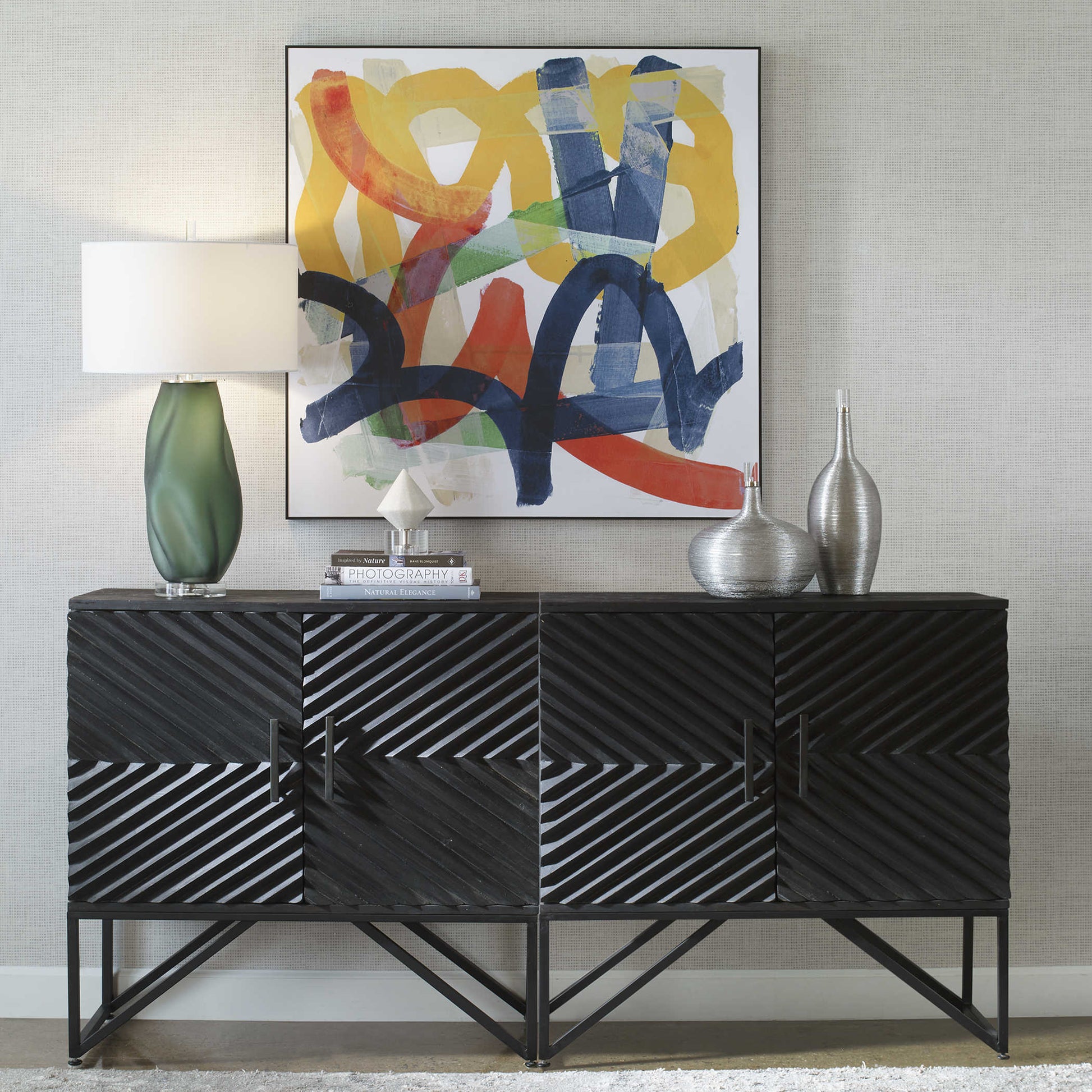 Straight up shot of a colorful and abstract piece of art behind a modern black console with an accent lamp and sculptures. 