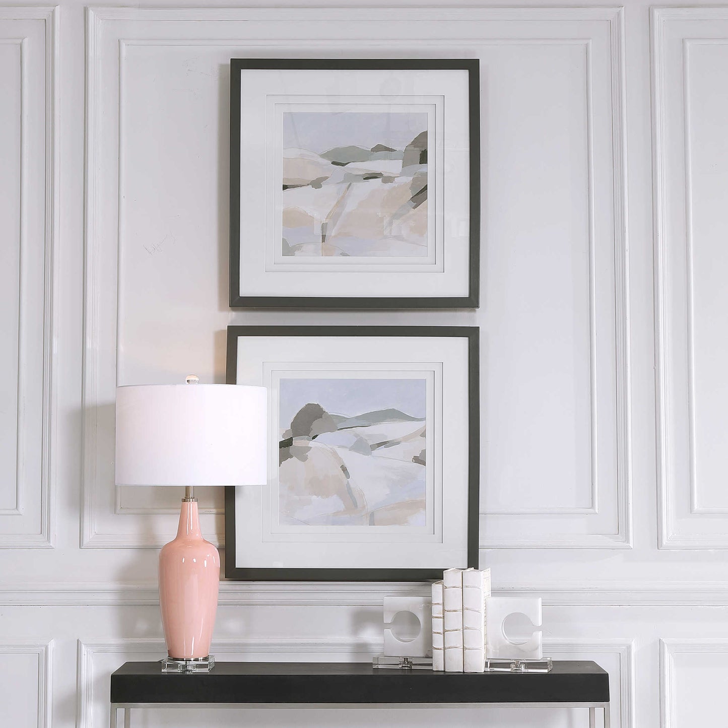 A pair of vertically stacked piece of green, beige, and sand landscapes in black frames above a modern, black console in front of of a pink lamp. 
