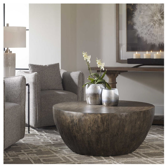 A room setting of round coffee table features mango wood veneer overlay in a a heavily textured aged walnut gray finish amongst occasional chairs. Console is against a wall with art above it in the background. The coffee table has a pair of vases on top with white orchids.