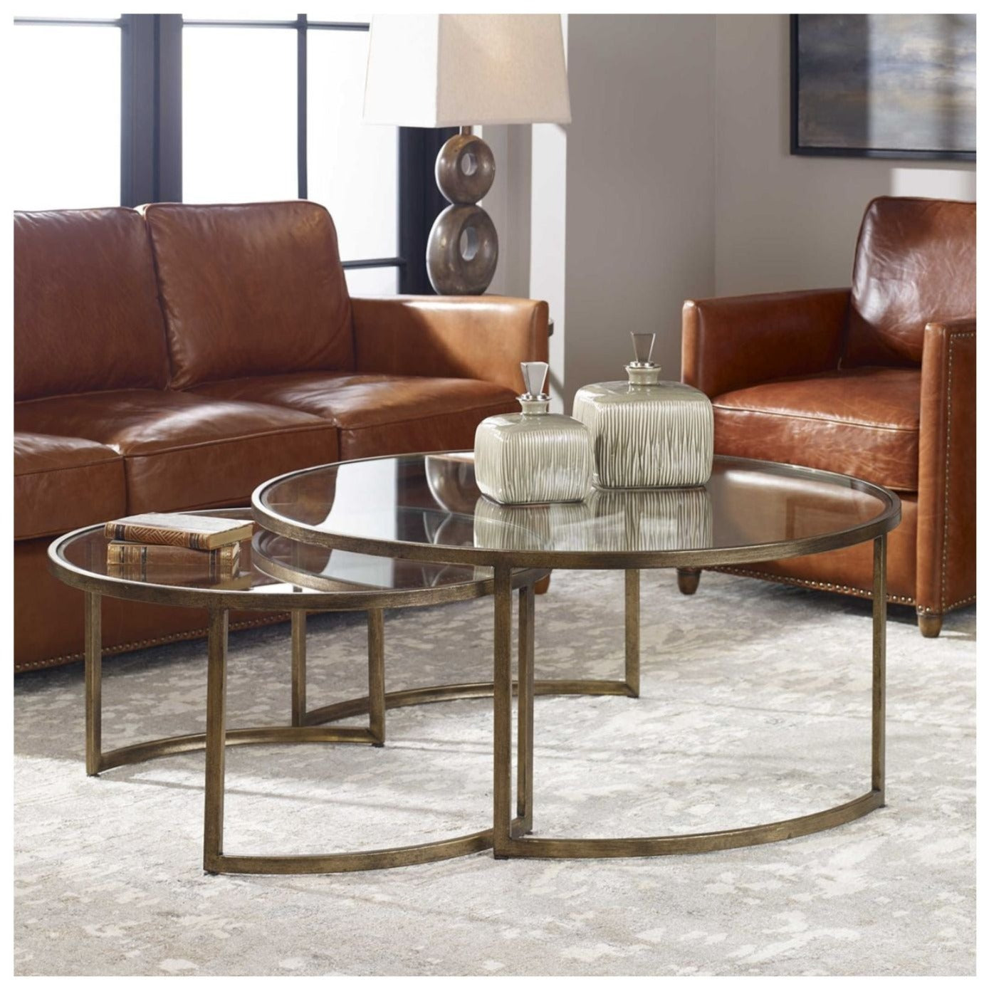 A room setting with leather chairs, a lamp, decorative boxes on top of the set of round gold frame and glass top nesting coffee tables.