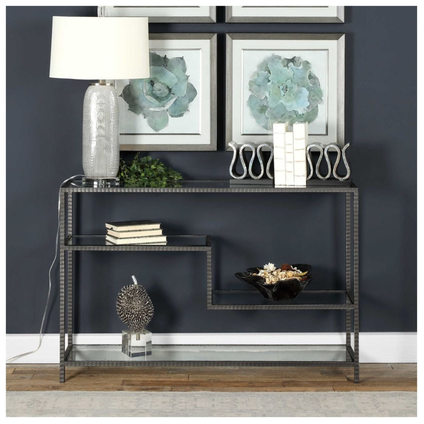 ribbed iron finished  console in an aged gunmetal with a light rust patina. Shelves are clear tempered glass decorated with a lamp, sculpture, books, bowl and book ends with overhead art..