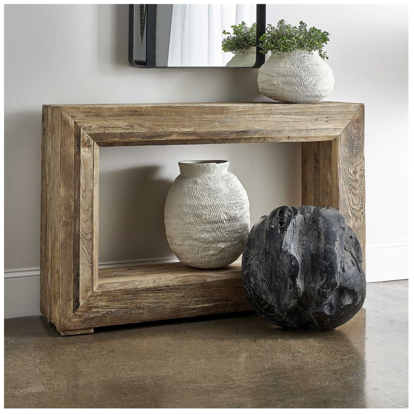 Room setting with console of character in rich reclaimed elm wood, featuring a beveled open center design. Decorated with large white vases on top and bottom, large wood ball sitting on floor in front and mirror hanging on wall above.carved 