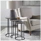 Set of three functional nesting tables constructed in an aged black iron, featuring a textured cast aluminum slab top finished in a plated antique nickel. In a room setting with a lamp on the larger table and a chair beside the set of three tables.