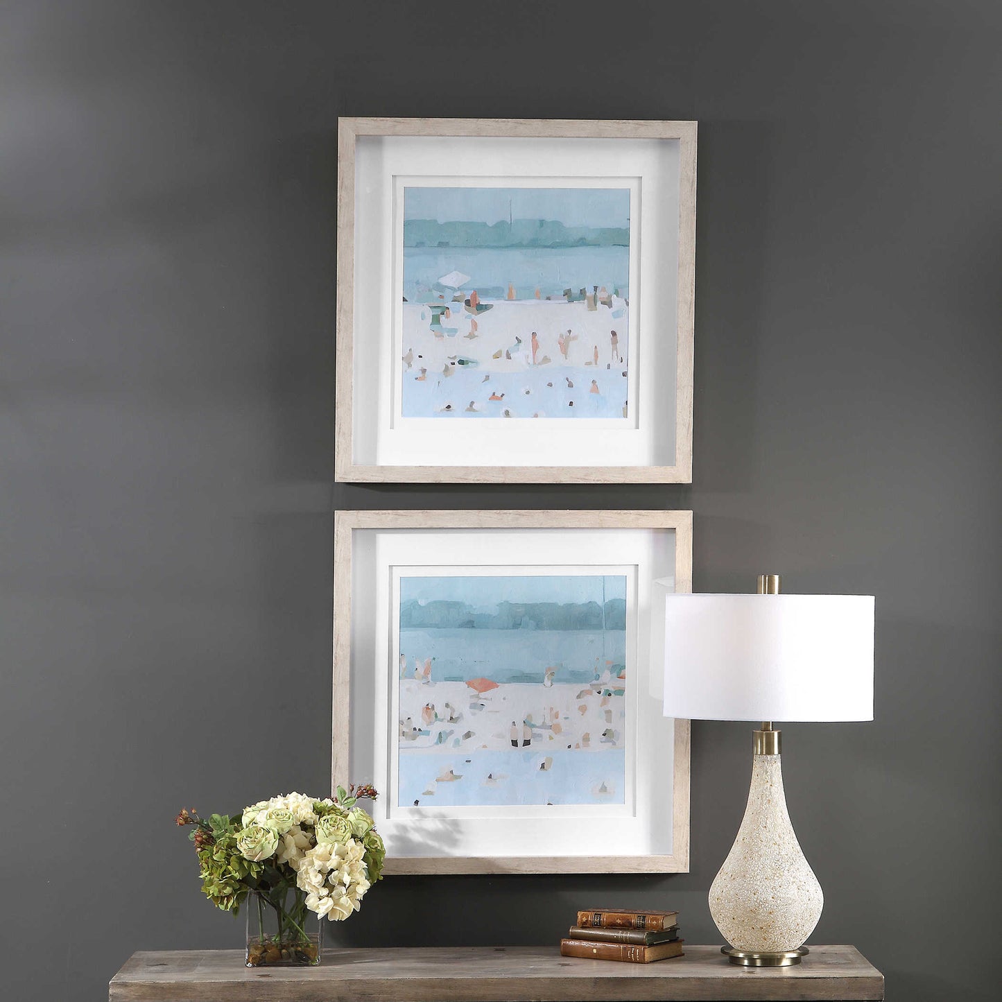 Stacked small art depicting a beach coastal scene on top of a console with a lamp, books, and flowers.