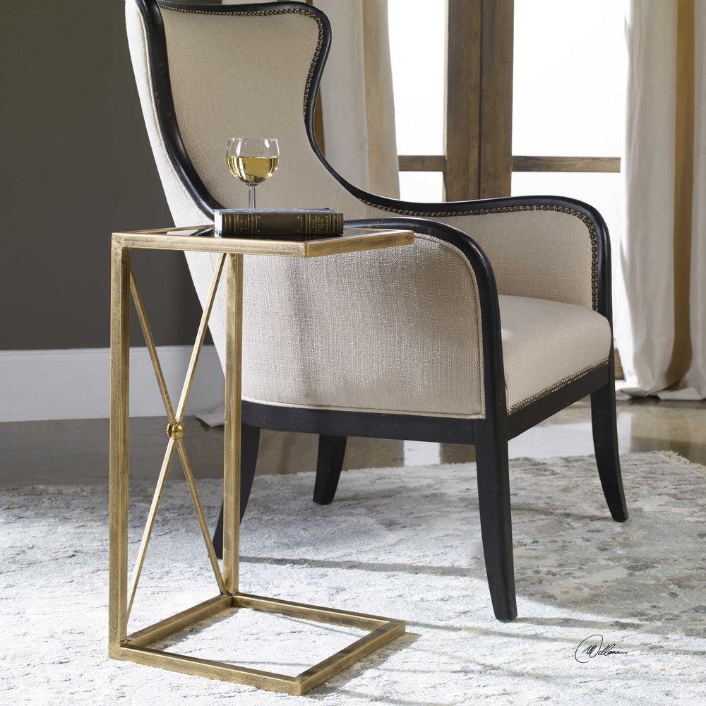A black and gold end table with a geometric metallic frame and a glass top, placed on a rug next to a chair.