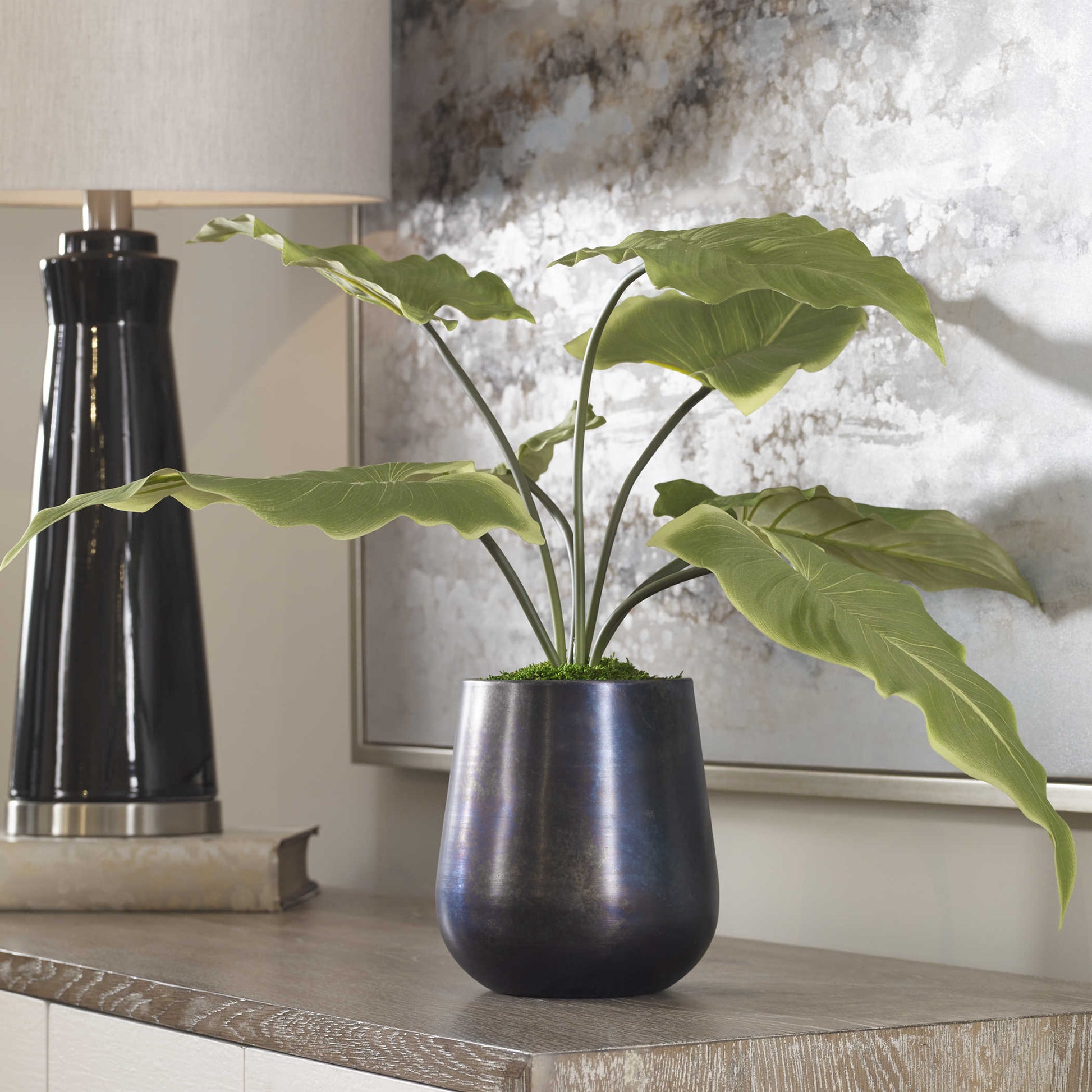 Fake plant and pot in oxidized planer in front of art on table surface.