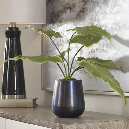 Fake plant and pot in oxidized planer in front of art on table surface.