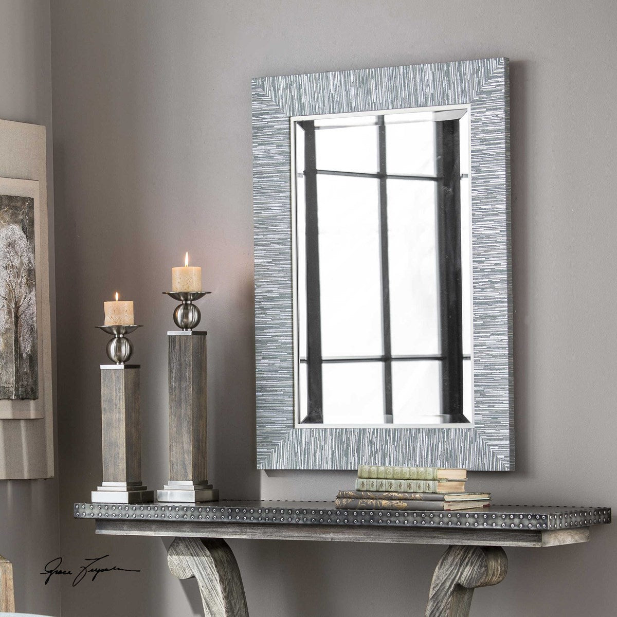 A rectangular blue-gray and silver striped mirror with a beveled edge and a silver leaf inner lip, mounted on a wall above a wooden console table with two candles and a reflection of a window in the background.
