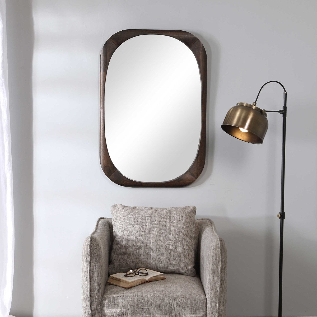 A rectangular, walnut-stained wood framed mirror with rounded edges hanging on a wall above a gray armchair, with a floor lamp beside the chair.