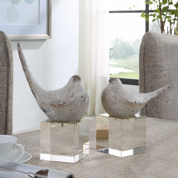A pair of silver-colored bird figurines with gray and rust distressed details, placed on top of a thick crystal block base.