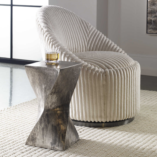 Silver twist accent table with a glass of whiskey on top next to a decorative chair on a textured white rug.