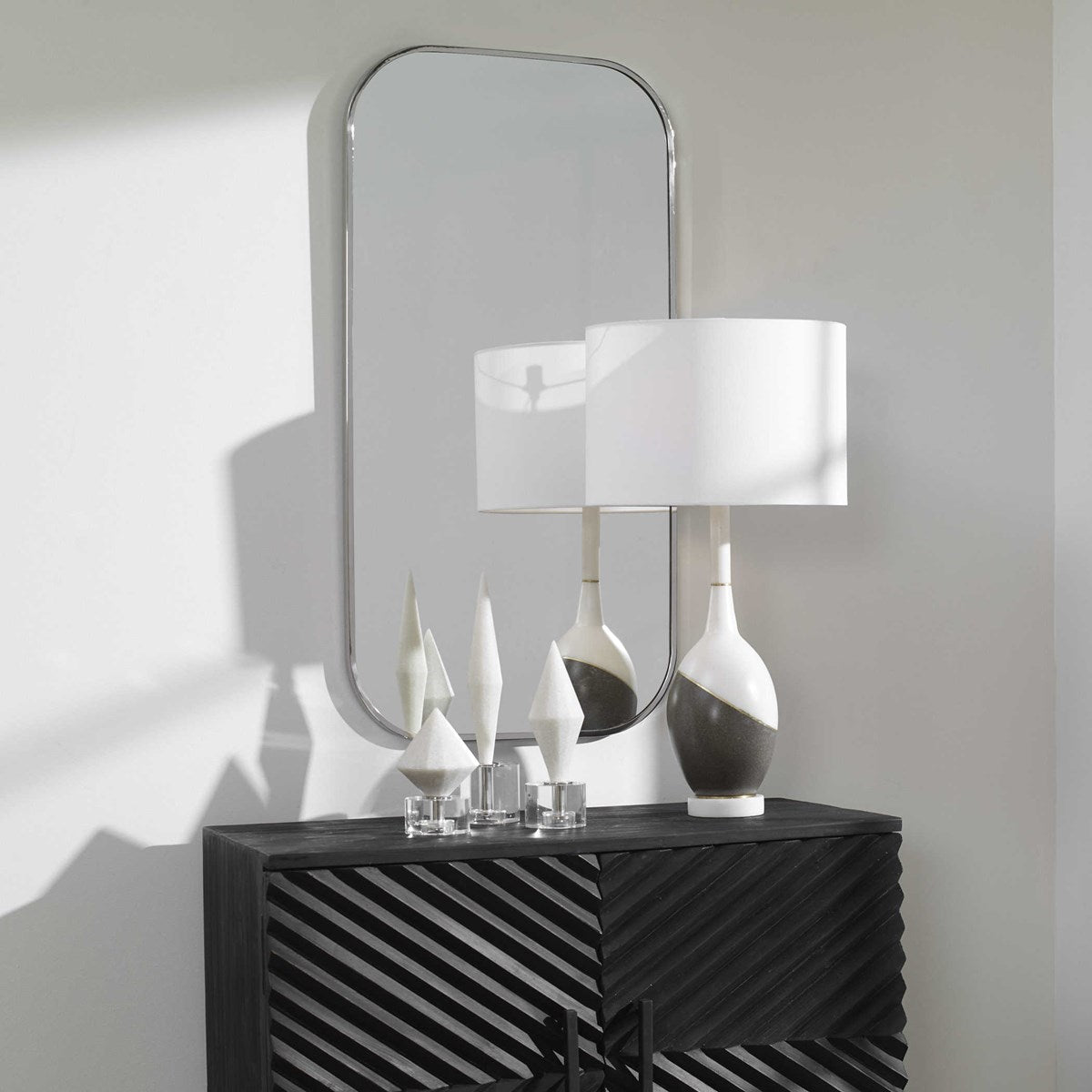 A polished nickel framed rectangle mirror mounted on a wall, reflecting a lamp and decorative objects on a cabinet.